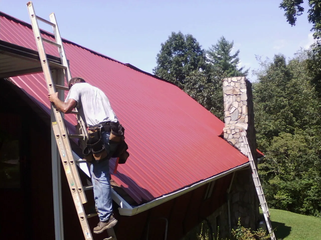 Roofer on a ladder beside a red metal roof and stone chimney — Soffit & Fascia Repair by Roofing Brothers in Germantown